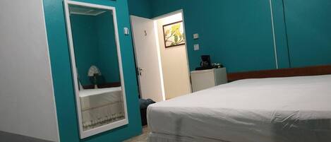 A room with teal walls, a bed, a TV on the wall, a fan, and a small kitchenette.