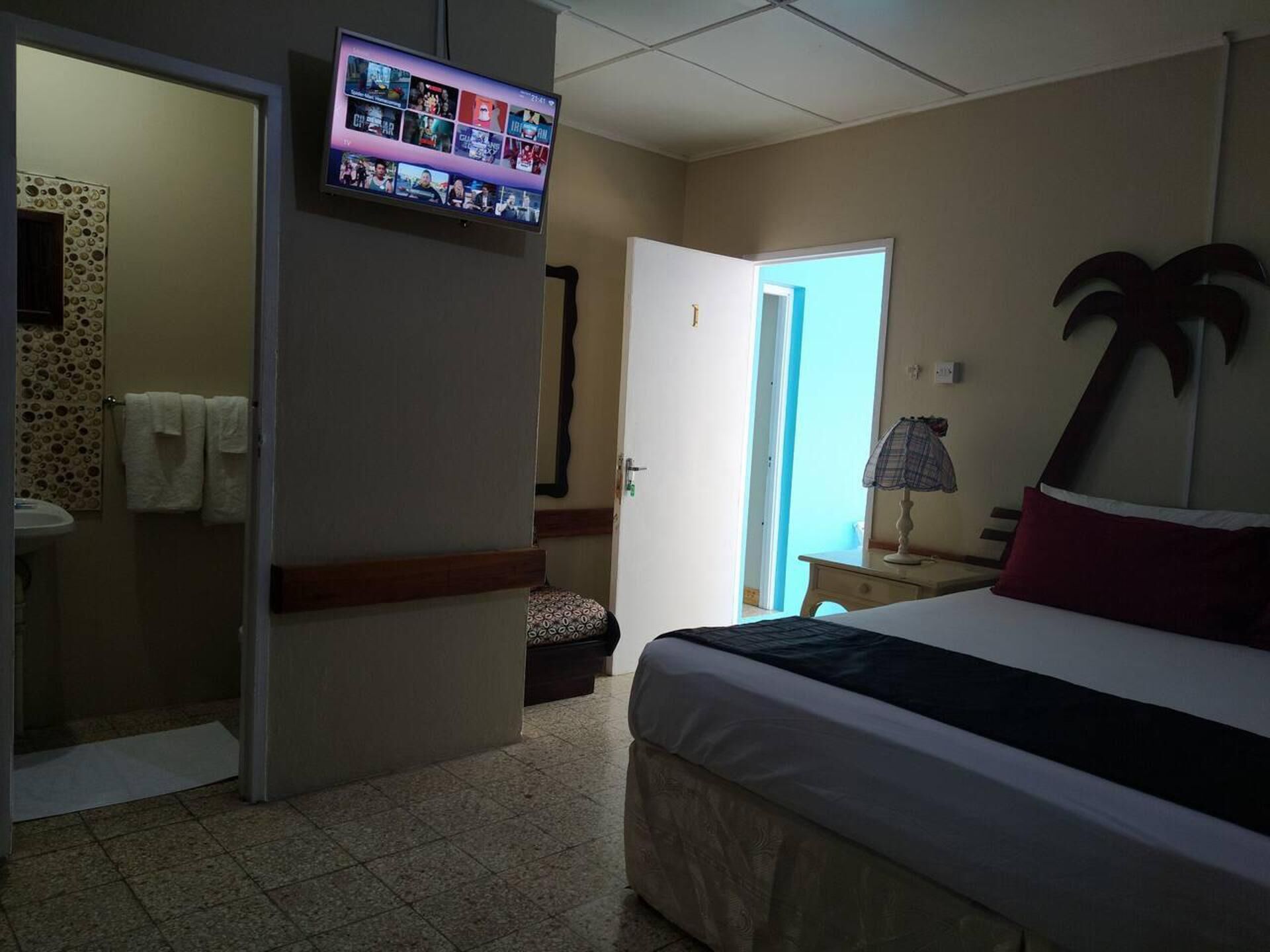 A hotel room with a bed, a television mounted on the wall, a bathroom with a towel rack, and a door leading to another room.