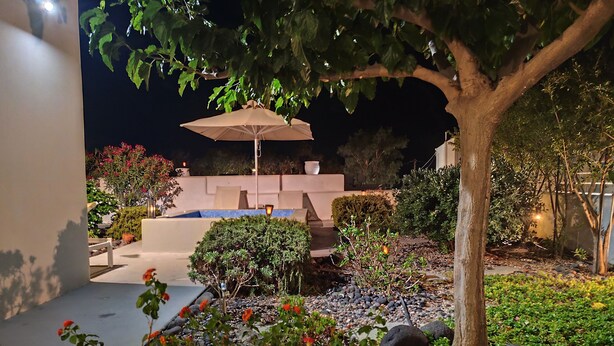 A well-lit outdoor area with a pool, lounge chairs, and a tree providing shade. - JB VILLA ,Stunning Santorini Escape w/heated jacuzzi and enclosed garden. (SANTORINI AIGEAN GREECE)