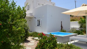 A white building with a swimming pool, surrounded by greenery and a beige umbrella. - JB VILLA ,Stunning Santorini Escape w/heated jacuzzi and enclosed garden. (SANTORINI AIGEAN GREECE)