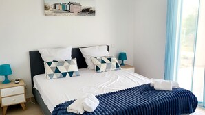 En pænt redt seng med et blåt tæppe og hvide puder, et lille natbord med en lampe og en vægmonteret aircondition.