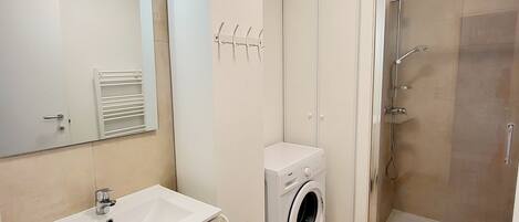 Un baño moderno con lavabo, espejo, lavadora y ducha con mampara de vidrio.