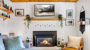 A cozy living room with a fireplace, a sofa, a chair, and a TV mounted above the fireplace. - Greer Cozy Cottage (Cabin 103) (Greer)