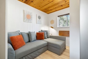 Uma sala de estar moderna com um sofá seccional cinza, almofadas laranja e cinza, teto de madeira e janela com vista para árvores.