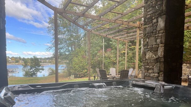 A hot tub area with a stone wall and wooden pergola, overlooking a lake and outdoor seating. - Lakehouse, Dock, Hot Tub & Tesla charger. Ideal for entertaining. Pet-Friendly! (Hiawassee)