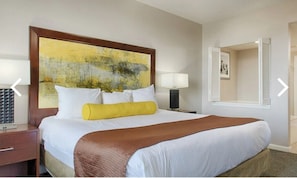A hotel room with a large bed, a headboard with a painting, a nightstand with a lamp, and a window with shutters. - Bike Week, 2027 March 5th through March 11th, 2027 ONLY (Daytona Beach)
