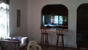 A living room with a sofa, a coffee table, and a vase with flowers. There is a kitchen area with a window and a ceiling fan. - Casa del campo (Tela)