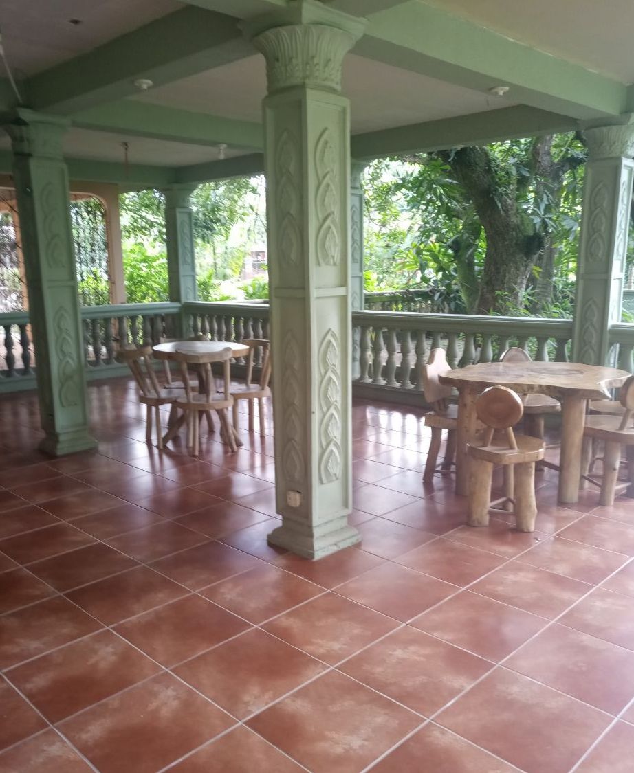 An outdoor covered area with tiled flooring, wooden tables and chairs, and decorative columns.