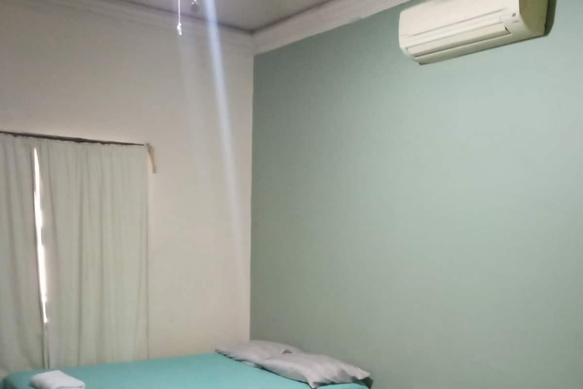 A room with two single beds, each with a white mattress and pillow, and a teal bedspread. There is a white air conditioning unit mounted on the wall above the beds.