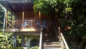 A wooden staircase leading to a covered upper level with a balcony, surrounded by lush greenery. - Casa del campo (Tela)