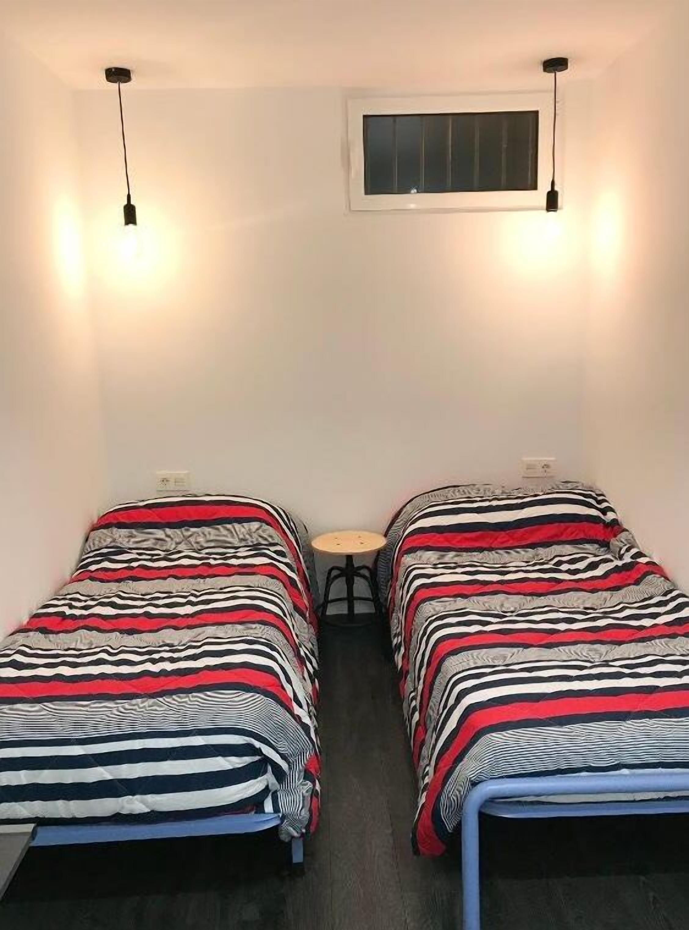 Una habitación pequeña con dos camas individuales, cada una con ropa de cama a rayas. Hay una mesita en el centro y una ventana sobre las camas.