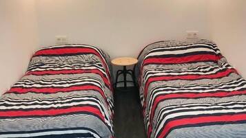 Una habitación pequeña con dos camas individuales, cada una con ropa de cama a rayas. Hay una mesita en el centro y una ventana sobre las camas.