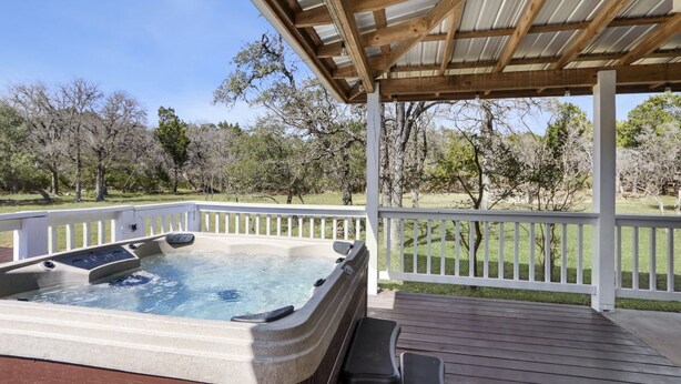 A hot tub on a wooden deck with a white railing, surrounded by trees. - Hot tub, sep. game room, wrap porch,quiet street,fire-pit,2 acres. All ages! (Wimberley)