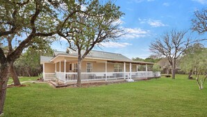 A two-story house with a covered porch, surrounded by a well-maintained lawn and trees. - Hot tub, sep. game room, wrap porch,quiet street,fire-pit,2 acres. All ages! (Wimberley)