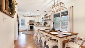 A modern kitchen with white cabinets, a wooden island, and a dining area with white chairs. - Hot tub, sep. game room, wrap porch,quiet street,fire-pit,2 acres. All ages! (Wimberley)