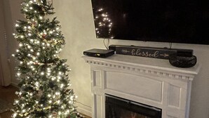 A living room with a Christmas tree, fireplace, flat-screen TV, and a mantel with a sign reading "Mercantile". - Hot tub, sep. game room, wrap porch,quiet street,fire-pit,2 acres. All ages! (Wimberley)