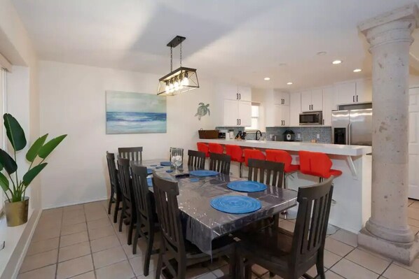 A dining area with a table set for six, surrounded by chairs. A kitchen with white cabinets and red chairs is visible in the background.