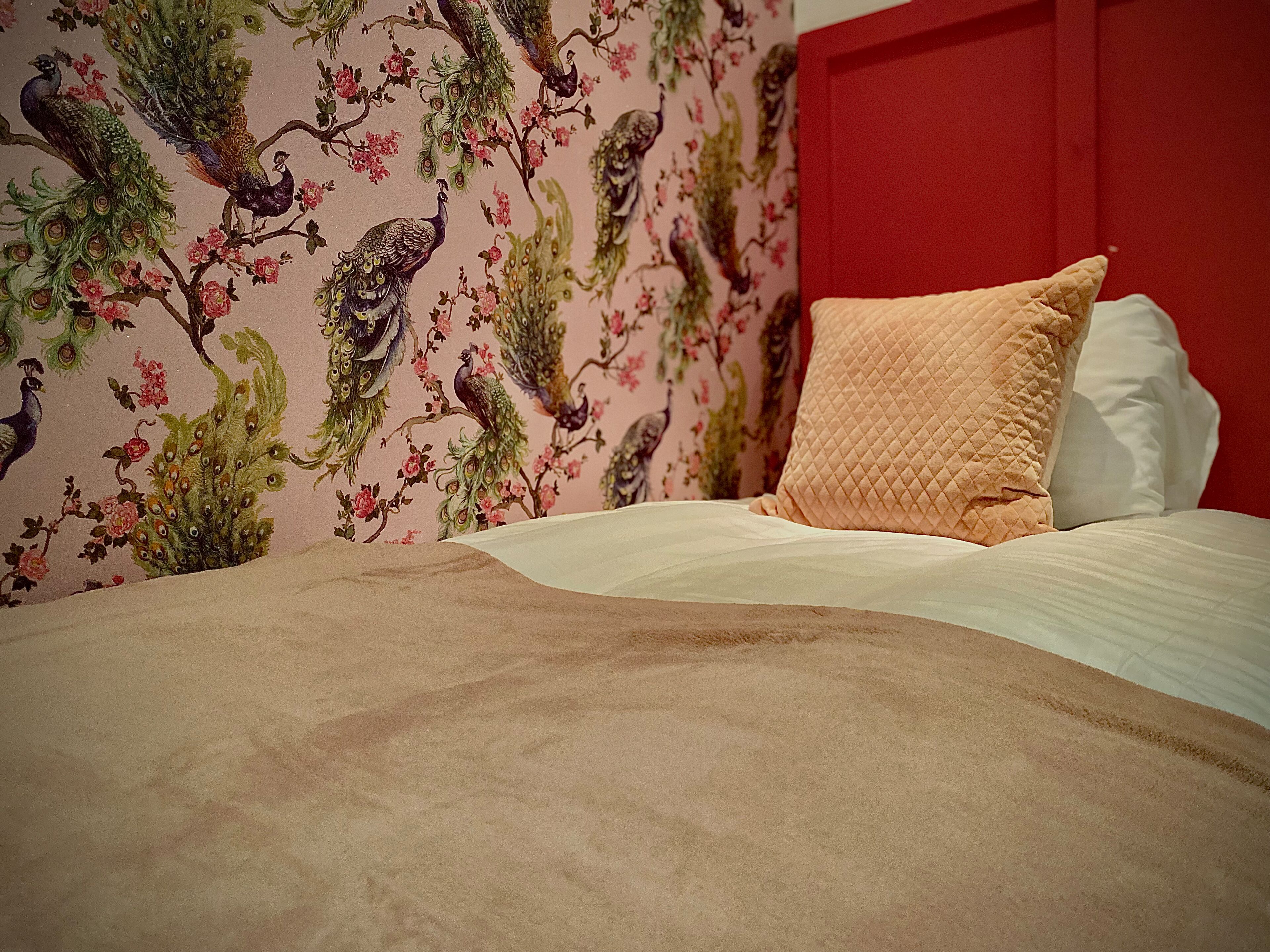 A bed with a quilted pillow, a red headboard, and a floral wallpaper with peacocks.