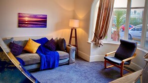 A living room with a sofa, a chair, a lamp, a painting, and a view of the street through a window.