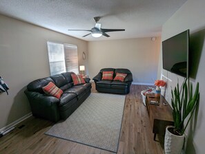 A living room with a black leather sofa, a smaller black sofa, a patterned rug, a ceiling fan, a TV, a wooden table with a vase of flowers, and a potted plant.