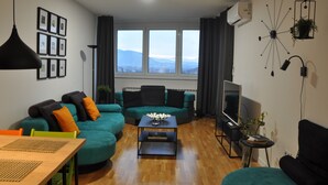 En moderne stue med en teal sofa, et træbord og et fjernsyn. Der er billeder i rammer på væggen og en plante på vindueskarmen.