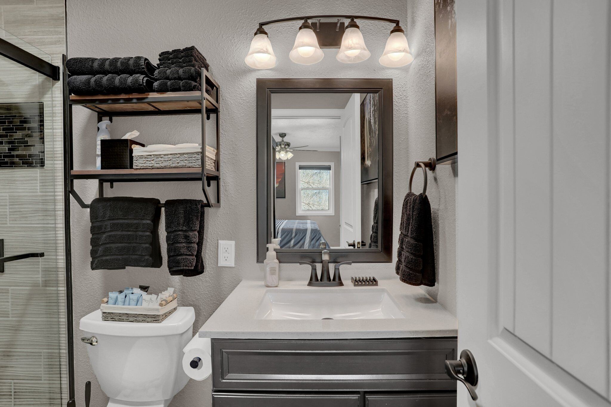 A bathroom with a white sink, a mirror, and shelves holding towels. There is a toilet with a basket of toiletries.