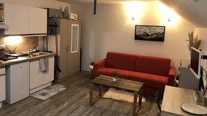 A compact living space with a kitchenette, a red sofa, a wooden coffee table, and a painting on the wall. - Lodging in the woods (Les Deserts)
