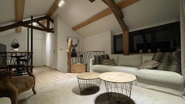 Uma sala de estar espaçosa com pé direito alto, vigas de madeira aparentes, um sofá grande, duas mesas redondas e uma área de jantar com mesa e cadeiras de madeira.