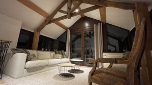 Una sala de estar acogedora con techo alto, un sofá, dos mesitas redondas y una silla de madera.