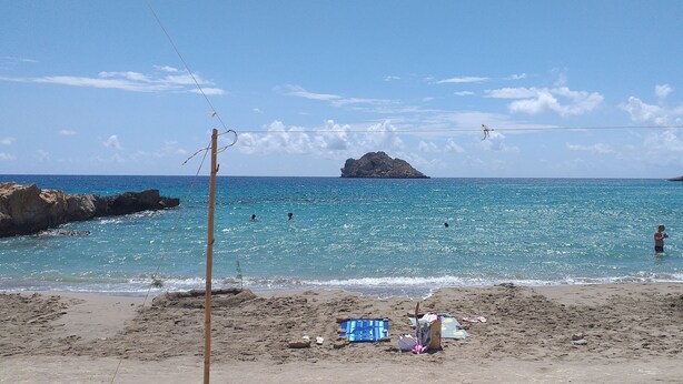 Une plage à l’eau d’un bleu limpide, avec un promontoire rocheux et quelques personnes qui se baignent.