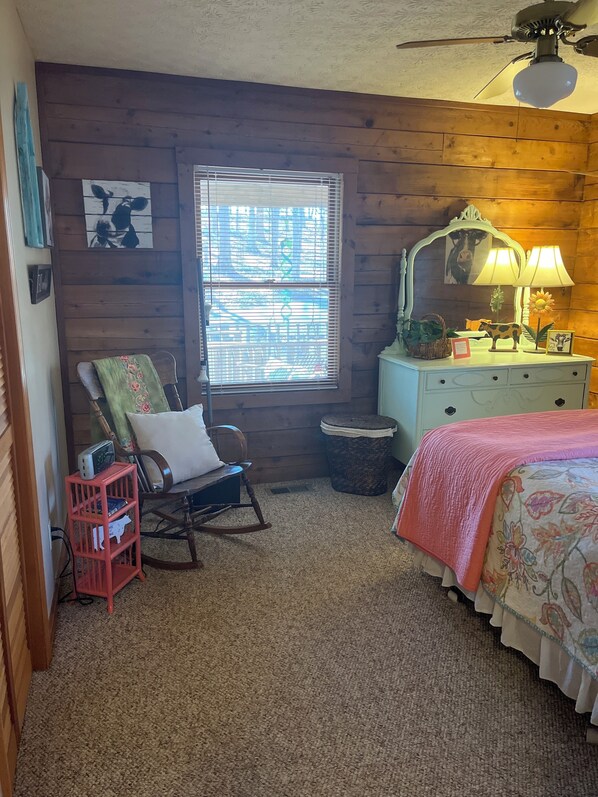 Une chambre confortable aux murs en bois, avec un fauteuil à bascule, un lit recouvert d’une courtepointe à motifs floraux, une commode avec un miroir et des lampes, et une fenêtre avec des stores.