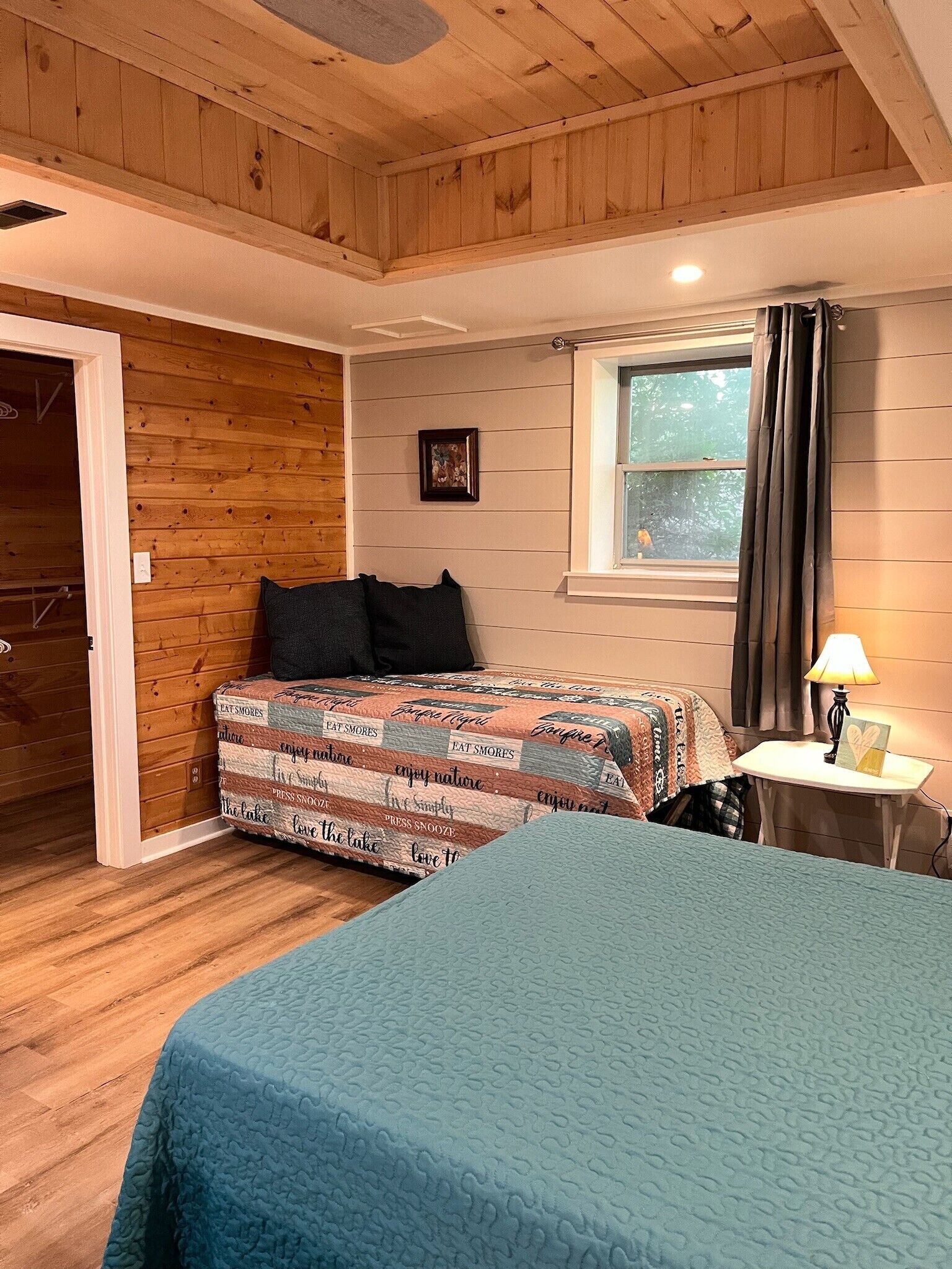 Une chambre avec un plafond en bois, un lit recouvert d’une courtepointe à motifs, une table de chevet avec une lampe et une fenêtre avec des rideaux.