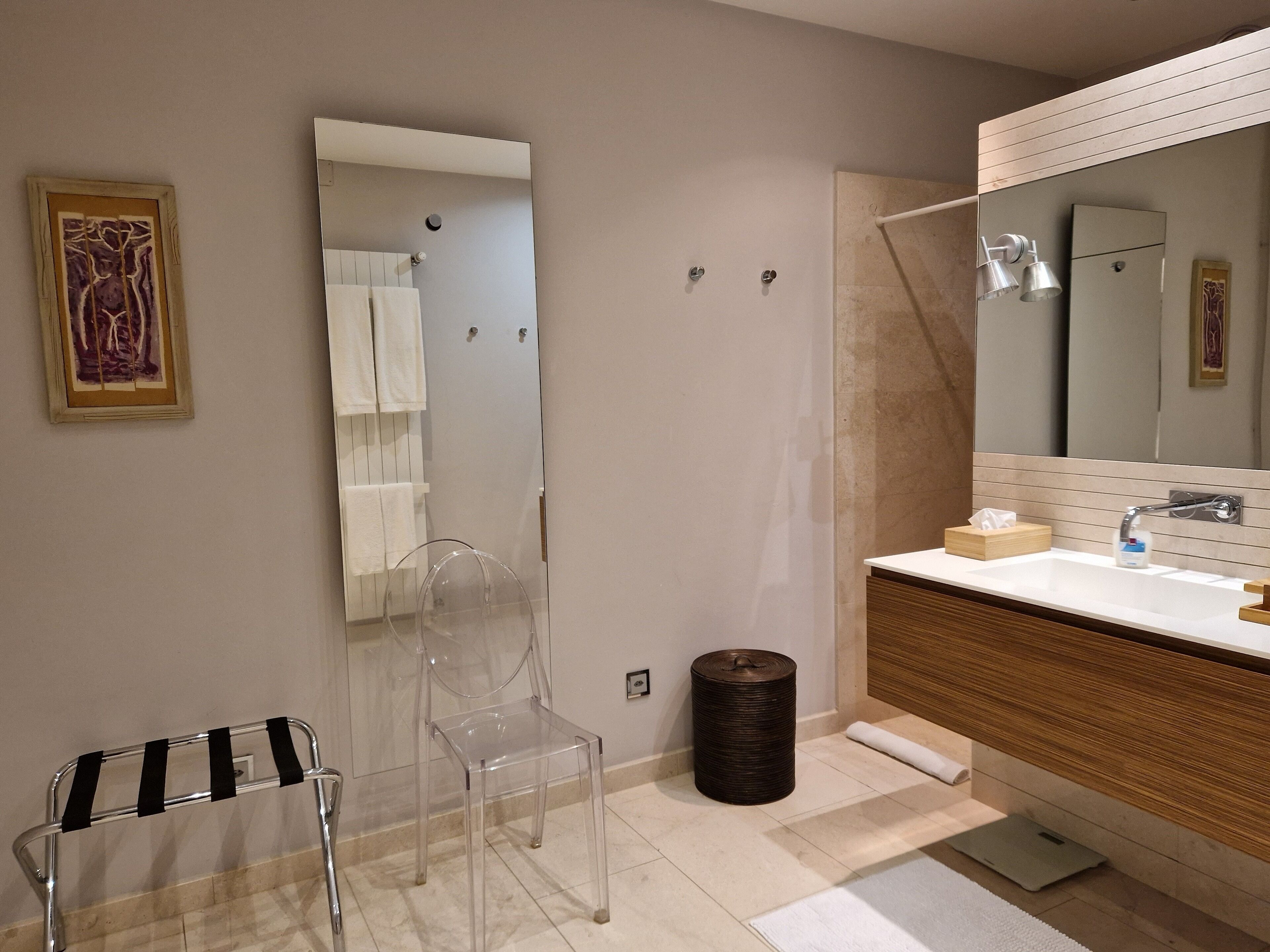 A modern bathroom with a large mirror, a sink with a wooden vanity, a clear chair, and a towel rack.