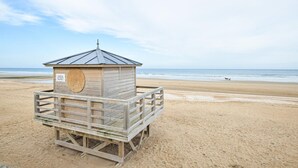 Une tour de surveillance en bois, agrémentée d’une pancarte, située sur une plage de sable avec l’océan en arrière-plan.