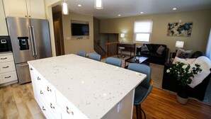 A modern kitchen with a large island, a dining area with chairs, a sofa, and a TV mounted on the wall.