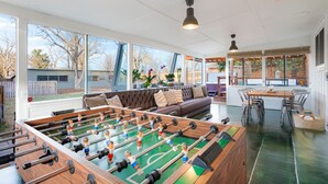 Ein geräumiger Raum mit einem Kicker, einer gemütlichen Couch, einem Esstisch mit Stühlen und großen Fenstern mit Blick ins Freie.