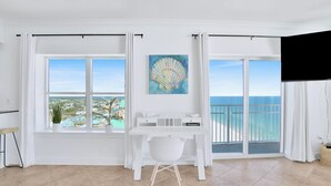 A bright room with a large window overlooking a coastal view, a white desk with a chair, and a painting of a shell on the wall. - Crystal Shores West 1401 | Guest Favorite! Gorgeous updates! Endless views! (Gulf Shores)