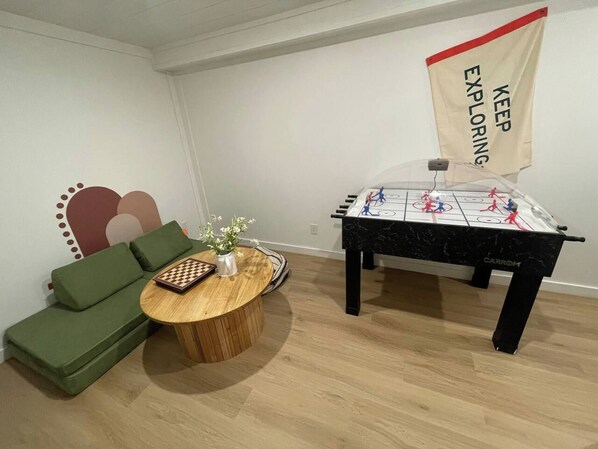 Una habitación con un sofá verde, una mesa de centro de madera con un tablero de ajedrez y una mesa de fútbolín.
