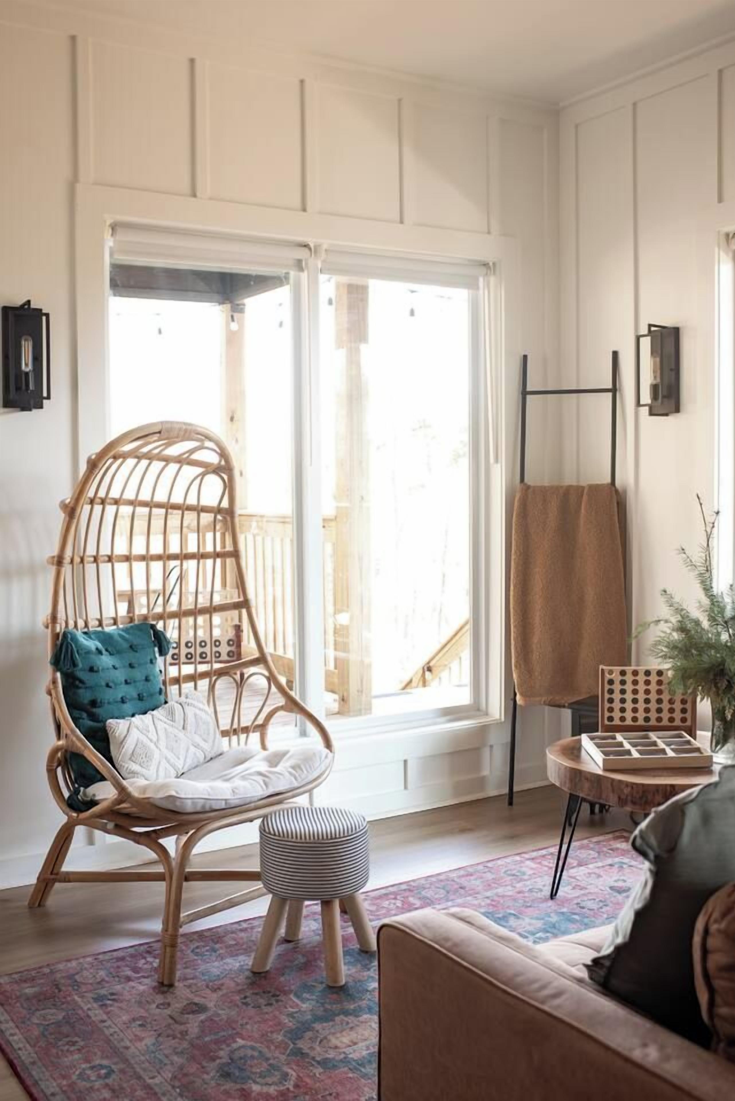 A room with a wicker chair, a small stool, a patterned rug, a wooden side table, and a view through a large window.