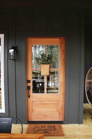 Una puerta de madera con un panel de vidrio, una cesta colgante con follaje y un felpudo con texto.