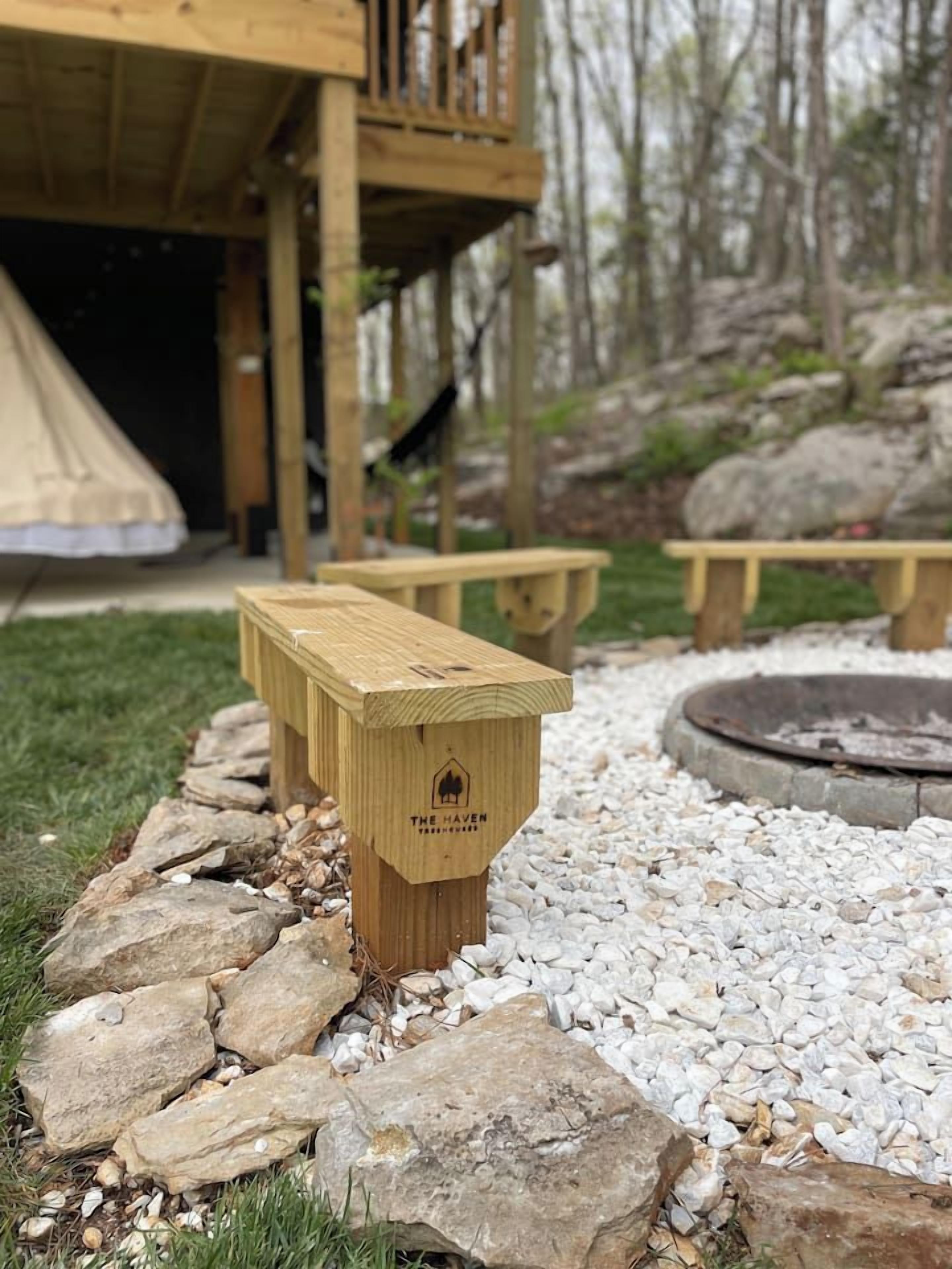 A wooden bench with a logo, surrounded by a stone border, in a garden area with a fire pit and a wooden structure in the background.
