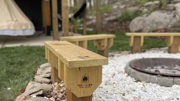 A wooden bench with a logo, surrounded by a stone border, in a garden area with a fire pit and a wooden structure in the background.