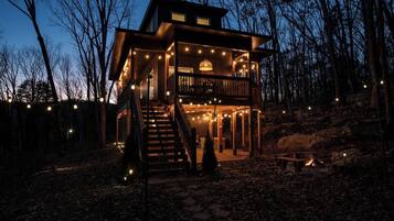 A two-story wooden cabin with a balcony, surrounded by trees and a stone pathway.