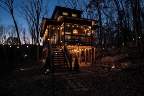 A two-story wooden cabin with a balcony, surrounded by trees and a stone pathway.