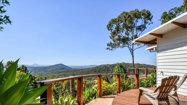 Une terrasse en bois offrant une vue sur des montagnes, un arbre et un bâtiment à la façade blanche.