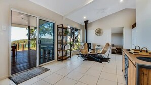 Un vaste espace de vie doté d’une grande porte-fenêtre coulissante donnant sur une terrasse extérieure, d’une bibliothèque en bois, de deux fauteuils en osier, d’un coin cuisine avec un plan de travail en bois et d’une horloge murale.