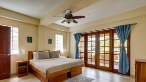 A bedroom with a bed, nightstands, a ceiling fan, a window with curtains, and a view through the door. - Beautiful Island Townhome Overlooking Caribbean (Placencia Village)