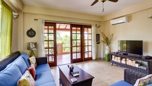 A living room with a blue sofa, a coffee table, a TV on a stand, and a ceiling fan. - Beautiful Island Townhome Overlooking Caribbean (Placencia Village)