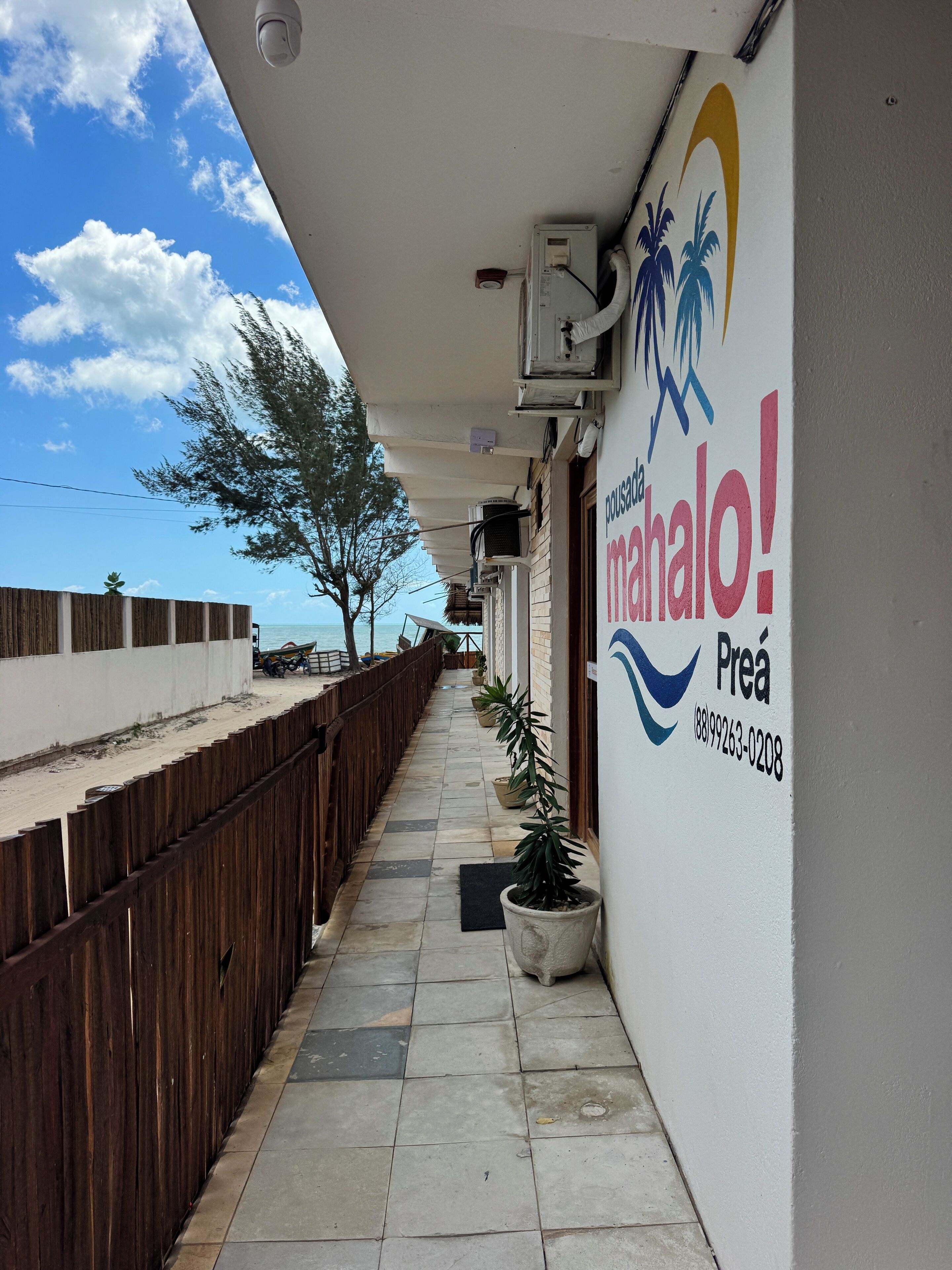 A corridor with a wooden fence, potted plants, and a sign for 'Manalo! Preá' with a contact number.