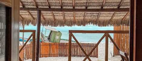 Una terraza de madera frente a la playa, con techo de paja, barandillas de madera y vista al océano.